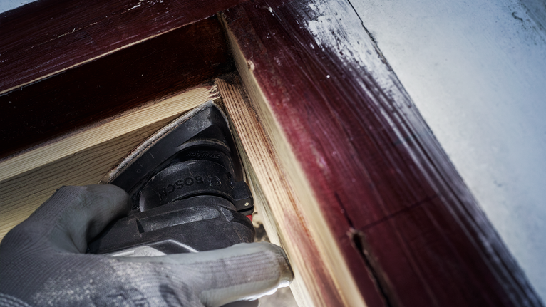 안전 장비를 착용한 사람이 페인트칠된 창틀 근처의 나무를 사포질하고 있습니다.