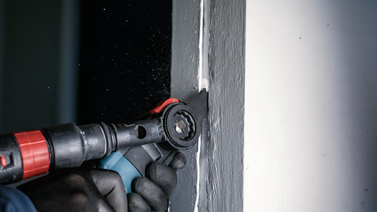 PRO MAII 32 SLC 멀티커터 날, 32 x 70mm 안전 장비를 착용한 사람이 진동하는 멀티툴을 사용하여 창문 근처의 석고를 자르고 있습니다.