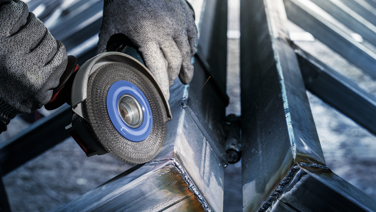 안전 장비를 착용한 사람이 각도 연삭기로 용접된 금속 빔을 연마하고 있습니다.