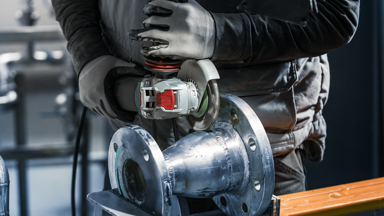 안전장비를 착용한 사람이 전동공구를 이용해 금속 파이프를 갈아내고 있습니다.