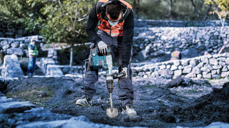 안전 장비를 착용한 사람이 회전 망치를 사용하여 야외에서 땅을 파고 있습니다.