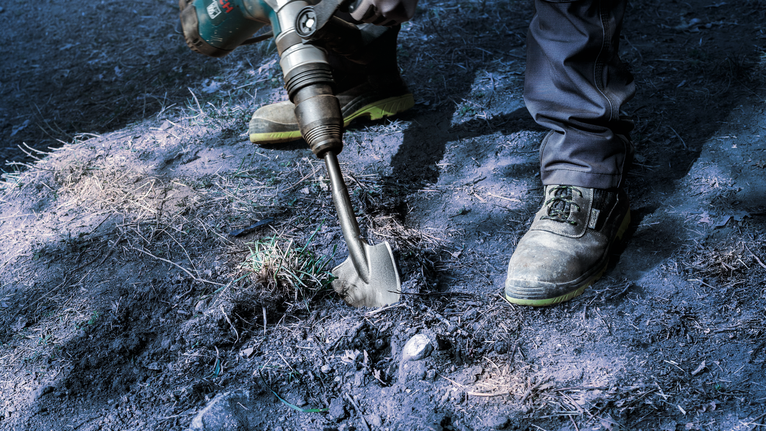 안전 장비를 착용한 사람이 끌이 달린 전동 공구를 사용하여 흙을 파헤칩니다.