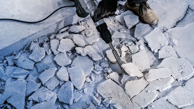 안전 장비를 착용한 사람이 철거 망치로 콘크리트 바닥을 부수는 모습.