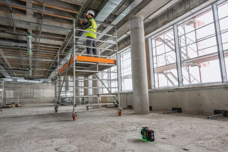 안전 장비를 착용한 사람이 비계 위에서 레이저 레벨링 도구로 선을 투사하며 작업하고 있습니다.