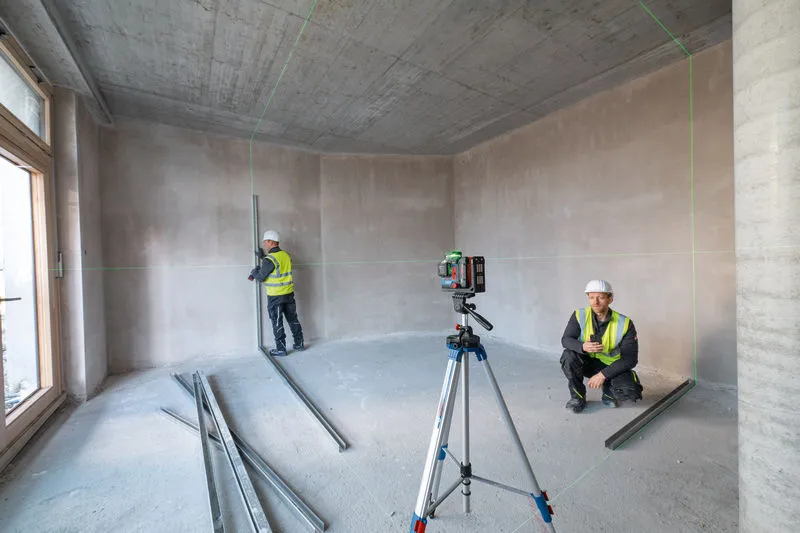 안전 장비를 착용한 두 사람이 미완성된 방에서 레이저 레벨링 도구를 사용하고 있습니다.