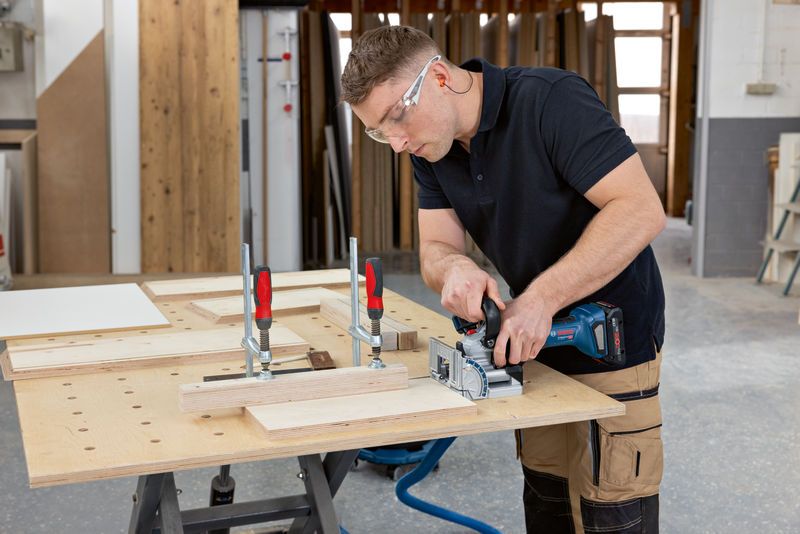 안전 장비를 착용한 사람이 클램프로 고정된 나무 조각에 비스킷 조인터를 사용하고 있습니다.