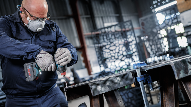 안전 장비를 착용한 사람이 산업 작업장에서 금속 빔에 구멍을 뚫고 있다.