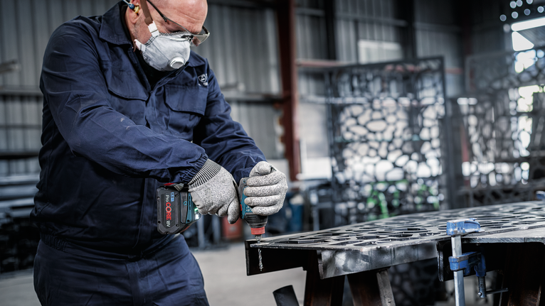 안전 장비를 착용한 사람이 작업장에서 금속판에 구멍을 뚫고 있습니다.