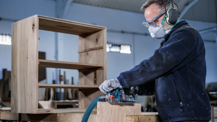 안전장비를 착용한 사람이 전동공구를 이용해 나무 가구를 연마하고 있습니다.