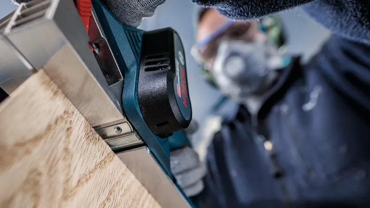 안전 장비를 착용한 사람이 나무판 위에서 전동 剪剪기를 사용하고 있습니다.