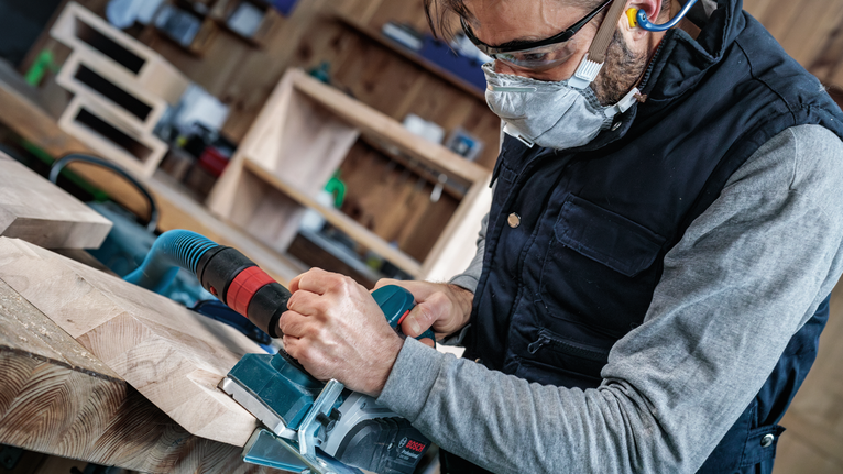 안전 장비를 착용한 사람이 전기톱을 조작하여 나무판을 자르고 있습니다.