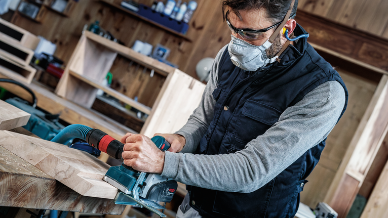 안전 장비를 착용한 사람이 전동 평면기를 사용하여 나무판을 매끄럽게 다듬고 있습니다.