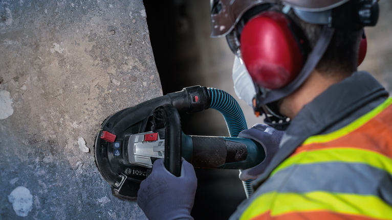 안전 장비를 착용한 작업자가 휴대용 전동 그라인더로 콘크리트를 닦고 있습니다.