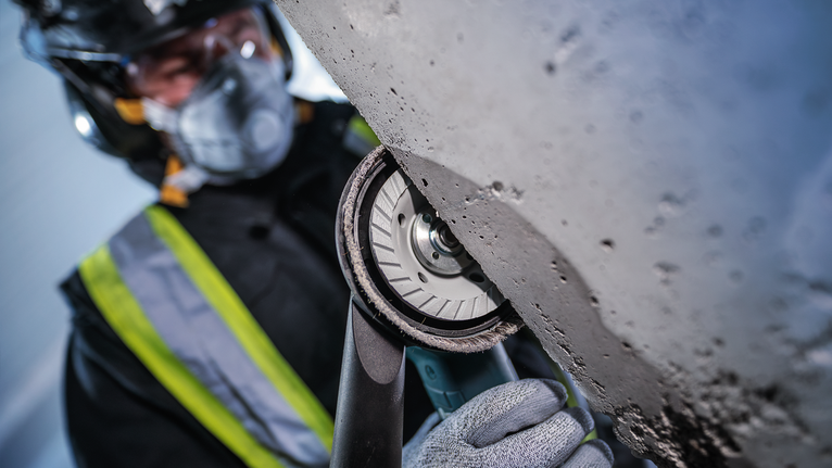 안전 장비를 착용한 사람이 휴대용 전동 공구를 이용해 콘크리트를 갈아내고 있습니다.