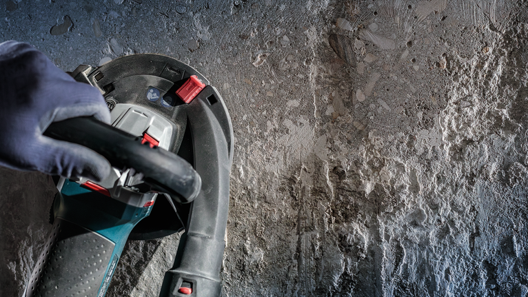 안전 장비를 착용한 사람이 거친 콘크리트 벽에서 전동 공구를 조작하고 있습니다.