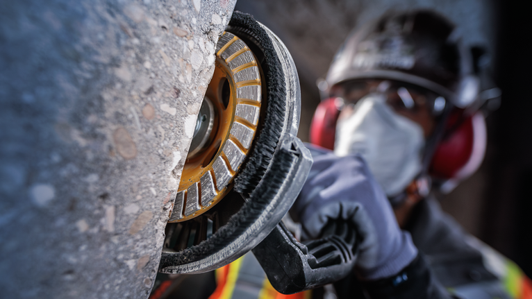 안전장비를 착용한 사람이 전동공구를 이용해 콘크리트 벽을 갈고 있습니다.