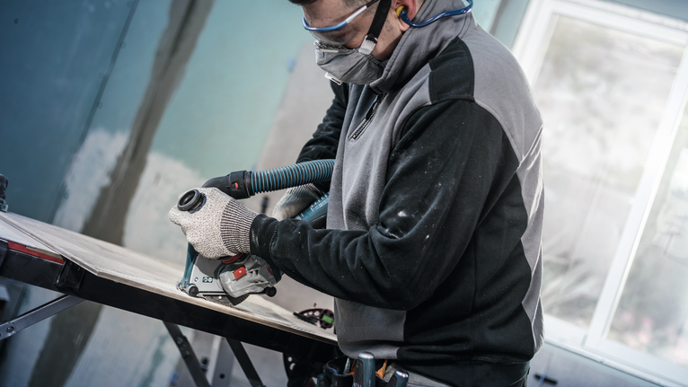 안전 장비를 착용한 사람이 진공 호스에 부착된 전동 공구로 나무를 다듬고 있습니다.