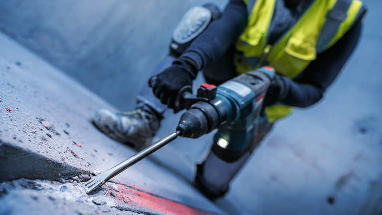 안전장비를 착용한 사람이 회전 망치로 콘크리트 표면을 깎고 있습니다.