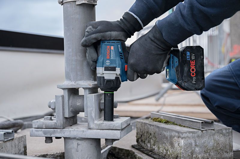 안전 장비를 착용한 사람이 무선 충격 렌치를 이용해 금속 구조물에 큰 볼트를 고정하고 있습니다.