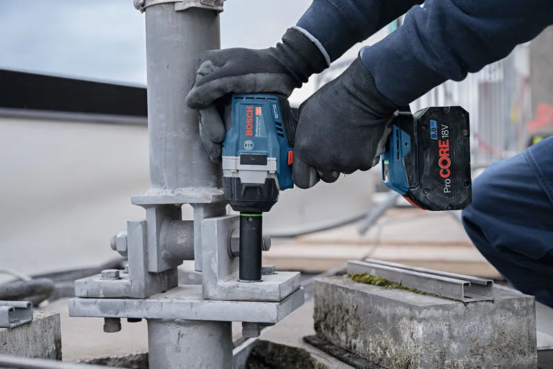 안전 장비를 착용한 사람이 무선 충격 렌치를 이용해 금속 구조물에 큰 볼트를 고정하고 있습니다.