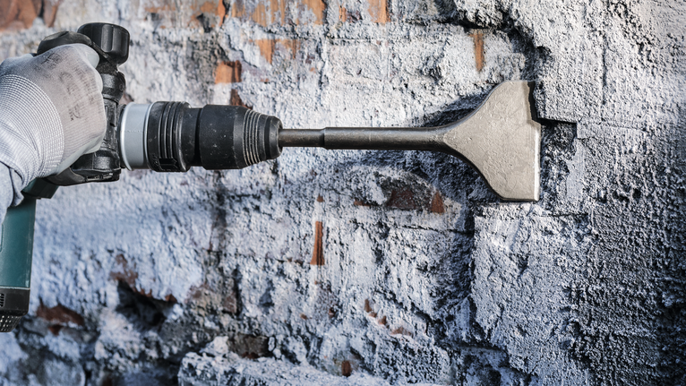 안전 장비를 착용한 사람이 끌 드릴을 사용하여 벽돌 벽에서 석고를 제거하고 있습니다.