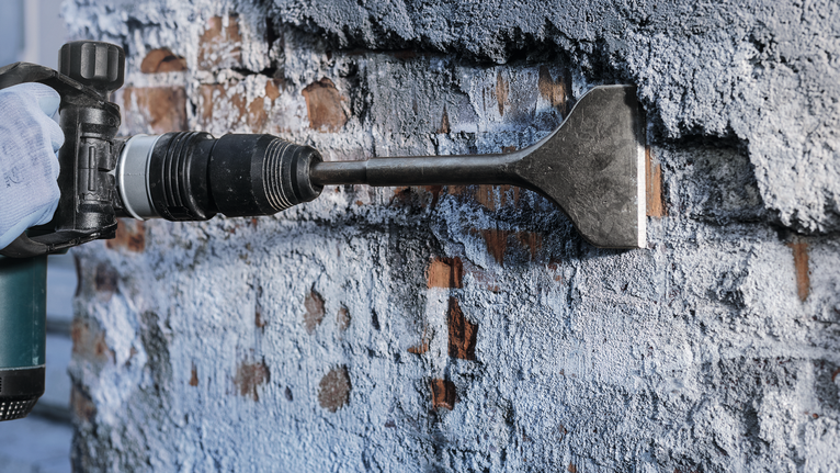 안전 장비를 착용한 사람이 끌 부착물을 사용하여 벽에서 석고를 제거하고 있습니다.