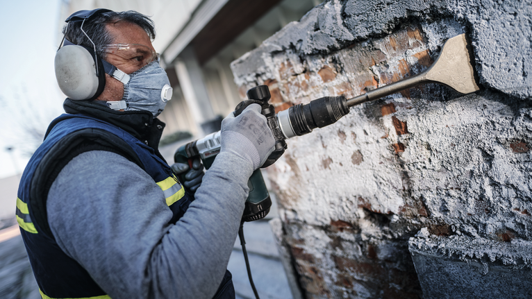 안전 장비를 착용한 사람이 철거 도구를 사용하여 벽에서 콘크리트를 제거하고 있습니다.
