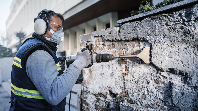 안전 장비를 착용한 사람이 전동 끌을 사용하여 벽돌 벽에서 모르타르를 제거하고 있습니다.