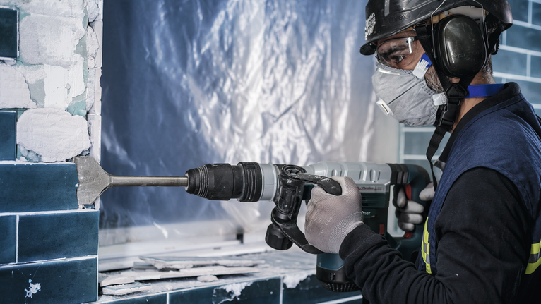 안전 장비를 착용한 사람이 회전 망치를 이용해 벽 타일을 제거하고 있습니다.