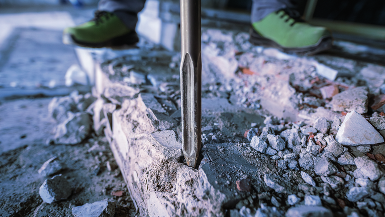 안전 장비를 착용한 사람이 철거 도구를 사용하여 콘크리트를 깨고 있습니다.