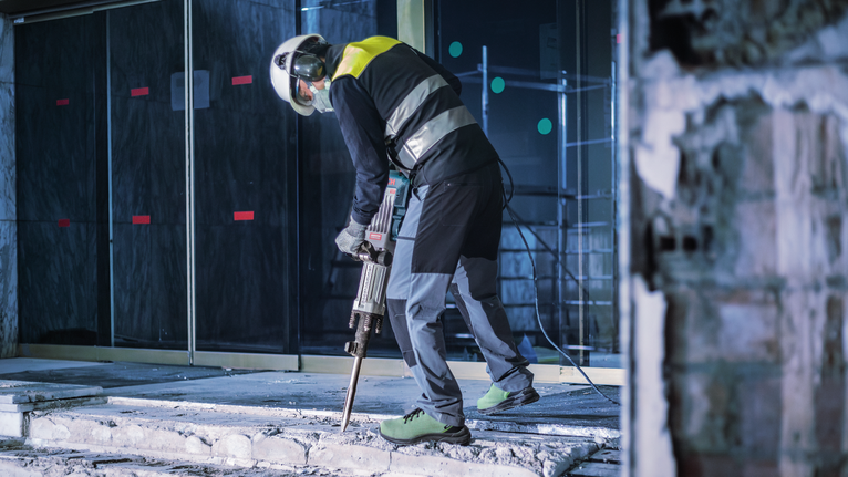 안전 장비를 착용한 사람이 철거 망치로 콘크리트를 부수는 모습.