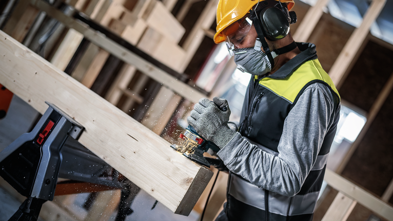 안전 장비를 착용한 사람이 전동 공구를 사용하여 나무 들보를 만들고 있습니다.