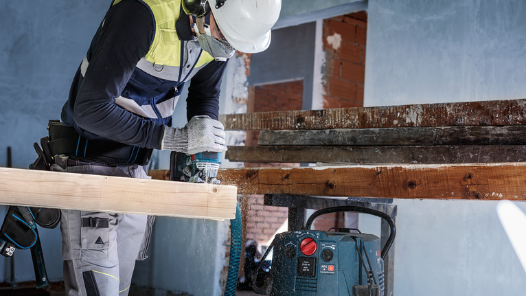 안전 장비를 착용한 사람이 건설 현장에서 나무 들보에 드릴을 박고 있습니다.