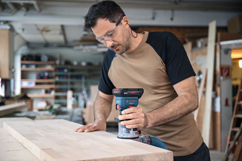 안전 장비를 착용한 사람이 무선 팜 라우터로 나무판의 가장자리를 다듬고 있습니다.