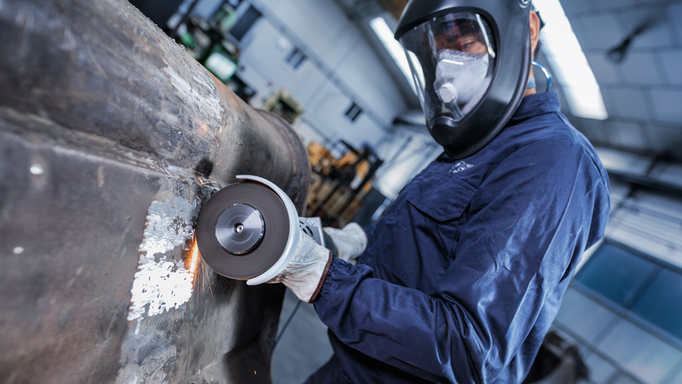 안전 장비를 착용한 사람이 산업 작업장에서 큰 금속 표면을 갈고 있습니다.
