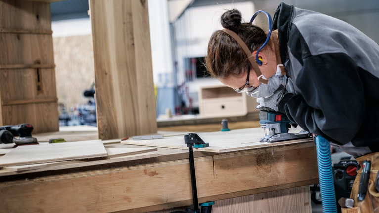 안전 장비를 착용한 사람이 나무판 위에서 목공 도구를 사용하고 있습니다.