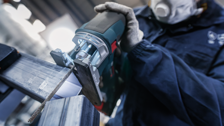 안전 장비를 착용한 사람이 왕복톱을 사용하여 금속 막대를 자릅니다.