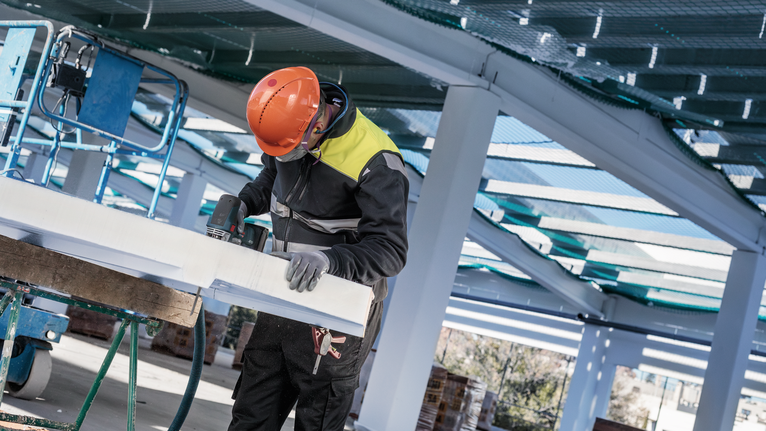 안전 장비를 착용한 사람이 건설 현장에서 큰 흰색 패널에 드릴을 박고 있습니다.