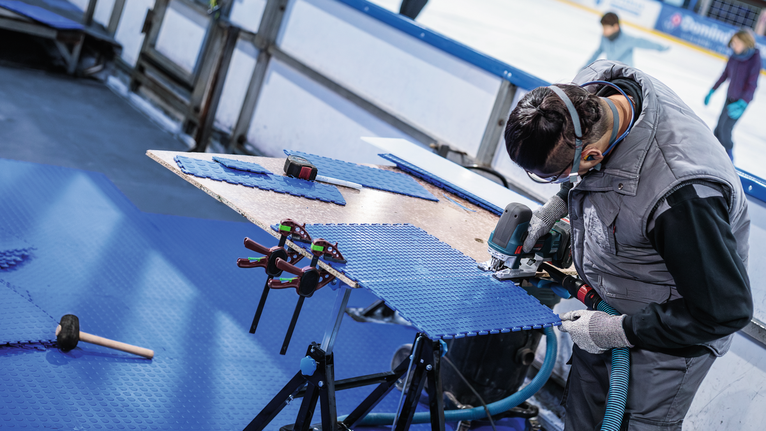 안전 장비를 착용한 사람이 전동 공구로 파란색 바닥 패널을 자르고 있습니다.