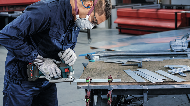 안전 장비를 착용한 사람이 작업대에서 전기톱으로 금속판을 자르고 있습니다.