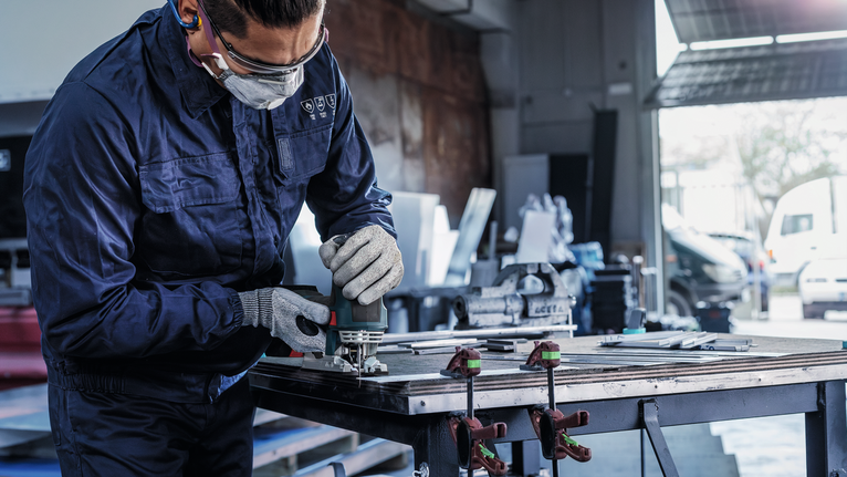 안전 장비를 착용한 사람이 작업대에서 전동 공구를 사용하여 금속을 연마하고 있습니다.