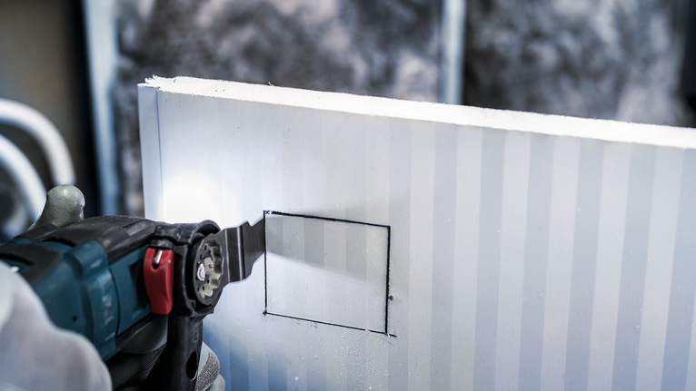 안전 장비를 착용한 사람이 전동 공구를 이용해 건식벽체에 정사각형 구멍을 자르고 있습니다.