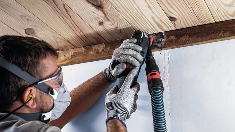 안전 장비를 착용한 사람이 전동 공구를 사용하여 나무 들보를 연마하고 있습니다.