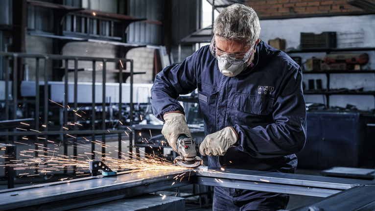 안전 장비를 착용한 작업자가 공장에서 금속을 갈아서 불꽃을 일으킨다.