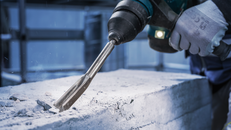 안전장비를 착용한 사람이 회전 망치로 콘크리트를 깎고 있습니다.