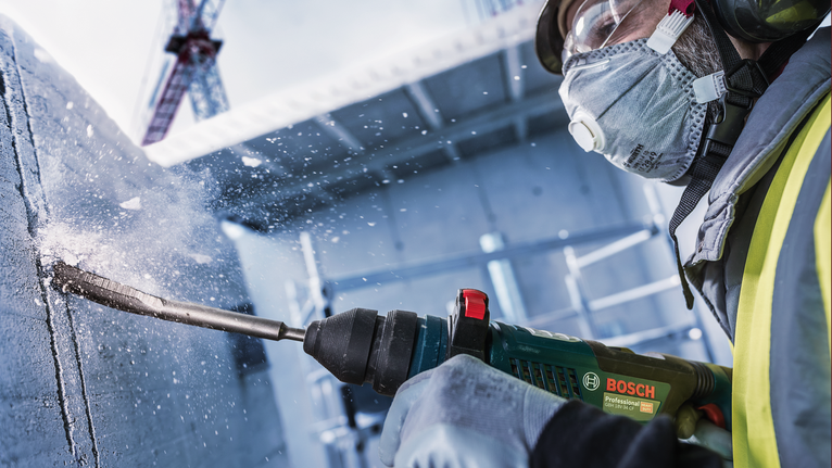 안전 장비를 착용한 사람이 콘크리트에 구멍을 뚫어 파편을 날리고 있습니다.