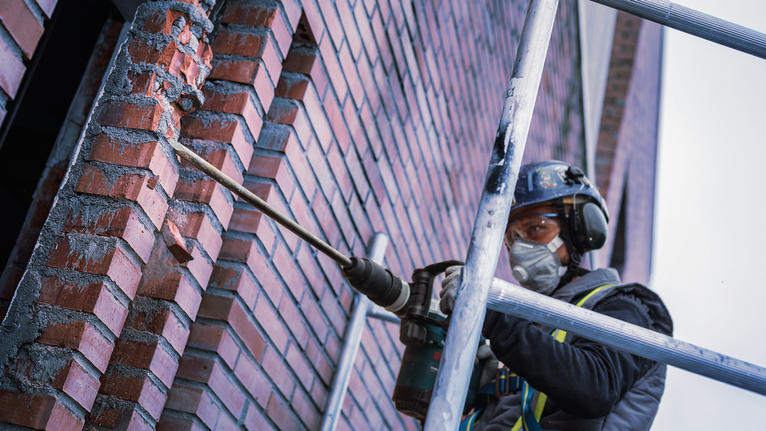 PRO SDS max-4C 포인트 치즐 600mm 안전장비를 착용한 사람이 비계 위의 벽돌 벽에 드릴을 박고 있습니다.