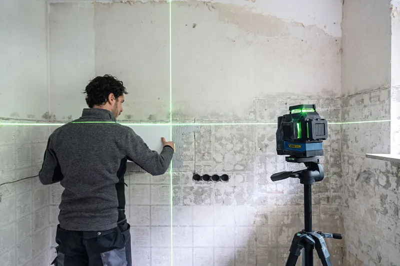 한 사람이 삼각대에 녹색 레이저 레벨링 도구를 올려놓고 벽에 타일을 정렬하고 있습니다.
