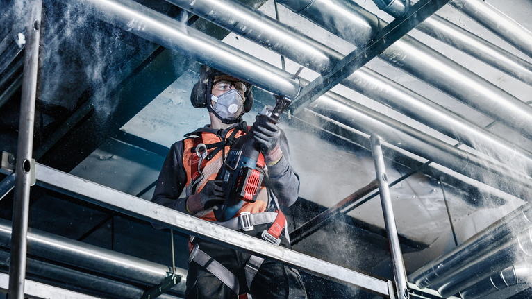 안전장비를 착용한 사람이 금속 천장 파이프에 구멍을 뚫고 있습니다.