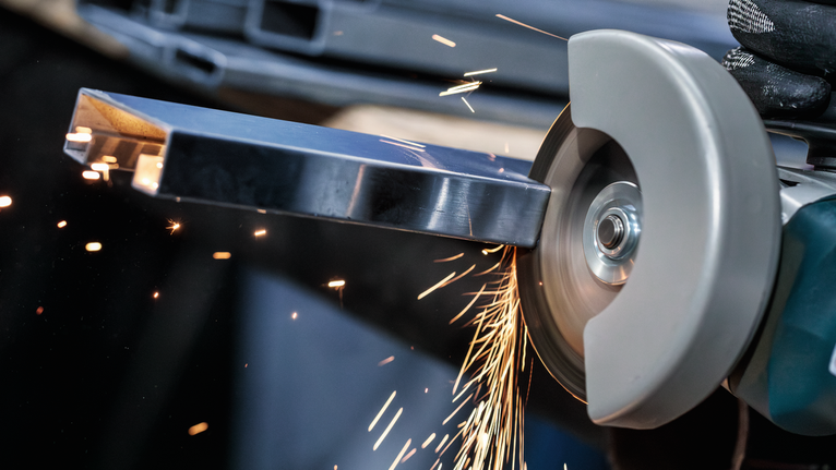 안전 장비를 착용한 사람이 손에 든 절단 도구로 금속 막대를 갈아내고 있습니다.